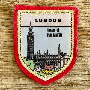 Op de afbeelding: Een schildvormige stoffen embleem met een rode vilten rand en gele zigzagstiksels. Het midden toont een gedrukte illustratie van het Londense parlementsgebouw en de Big Ben, met de tekst LONDON en Houses of PARLIAMENT erboven.