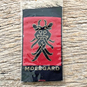 Puede incluir: Un parche rectangular con borde negro y centro rojo. El centro presenta un diseño negro estilizado con detalles dorados y la palabra "MOESGARD" en letras doradas. La palabra "MENOR" está debajo.