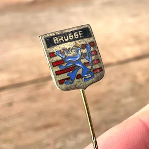 Op de afbeelding: Vintage speld met het woord "BRUGGE" in witte letters op een zwarte achtergrond. De speld heeft een schildvorm met een blauwe leeuw en rode strepen. De speld is goudkleurig.
