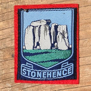 May include: A rectangular patch with a red border features an embroidered image of Stonehenge. The patch has a blue background with the monument in white and a green base. The word "STONEHENGE" is displayed in white letters.