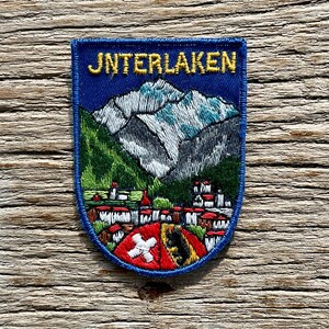 Op de afbeelding: Geborduurde patch met een blauwe rand en achtergrond. De patch toont het woord "JNTERLAKEN" in gouden letters boven een bergketen met groene en grijze details. Onder de bergen bevindt zich een stad met rode daken en een schild met een wit kruis.