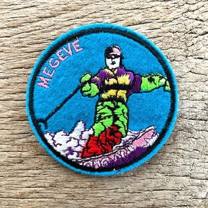 Op de afbeelding: Een ronde, geborduurde patch met een skiër in actie tegen een turquoise blauwe achtergrond. De skiër draagt een paars en groen outfit en skiet op een besneeuwde helling. Het woord "MEGEVE" is roze geborduurd langs de rand.