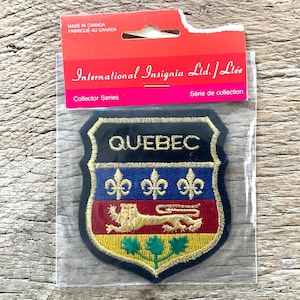 Op de afbeelding: Een Quebec-patch met een schildontwerp. De patch heeft een zwarte rand met gouden afwerking, het woord "QUEBEC" in goud en symbolen van leeuwen en fleur-de-lis. De patch zit in een doorzichtige plastic verpakking met een rood en roze label.