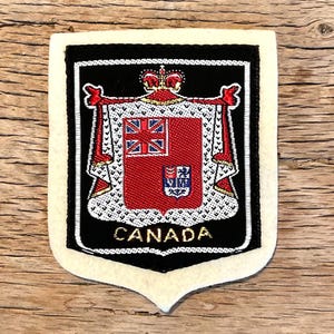 Op de afbeelding: Een stoffen patch met het Canadese wapen. De patch heeft een zwarte rand en een crèmekleurige rand. Het wapen is rood met een Union Jack en een kleiner embleem. Het woord "CANADA" is in goud geborduurd.
