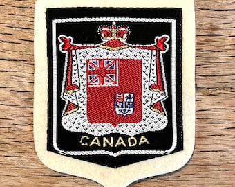 Puede incluir: Parche de tela con el escudo de armas canadiense. El parche tiene un borde negro y un borde color crema. El escudo de armas es rojo con una Union Jack y un emblema más pequeño. La palabra "CANADA" está bordada en oro.