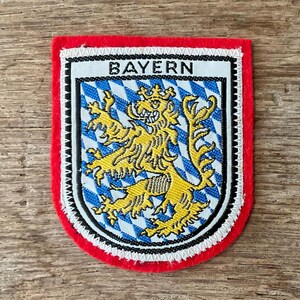 Peut inclure: Écusson brodé avec une bordure rouge et un bord cousu blanc. Le patch présente le mot "BAYERN" au-dessus d'un lion jaune sur un fond à motifs de losanges bleus et blancs.