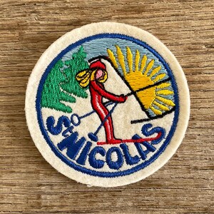 Op de afbeelding: Een ronde geborduurde patch met een skiër in een rood pak op een lichtblauwe achtergrond. Het ontwerp bevat groene dennenbomen, een heldere gele zon en de blauwe tekst ST NICOLAS gebogen langs de onderrand van de cirkelvormige rand.
