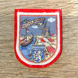 Op de afbeelding: Een rechthoekige patch met een rode rand en een gedetailleerd geborduurd ontwerp. Het ontwerp toont een molen, een boot en het woord "HOLLAND" op een banner. De onderkant van de patch heeft het woord "NEDERLAND" en een wapen.