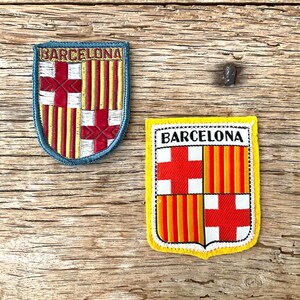 Op de afbeelding: Twee geborduurde patches met het embleem van FC Barcelona. Eén patch heeft een blauwe rand en het woord "BARCELONA" bovenaan. De andere heeft een gele rand en het woord "BARCELONA" bovenaan. Beide patches tonen de rood-gele kleuren van het team.