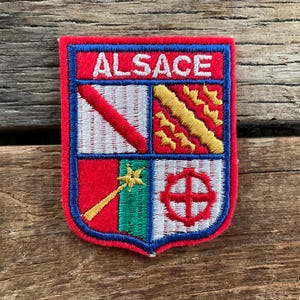 May include: A rectangular embroidered patch with a red border and the word ALSACE in white capital letters. The shield design is divided into four quadrants featuring a red diagonal stripe on white, a gold diagonal stripe, a gold star, and a red cross.
