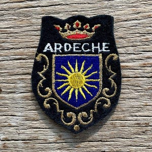 Op de afbeelding: Geborduurde patch met het woord "ARDECHE" in het wit, boven een blauw schild met een gele zonnestraal. De patch heeft een zwarte achtergrond met gouden decoratieve krullen en een rood-gouden kroon.