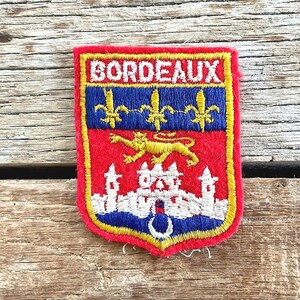 May include: Embroidered patch with the word "BORDEAUX" at the top. The patch features a red background with a blue stripe and gold fleur-de-lis. A gold lion and a white castle are also featured.