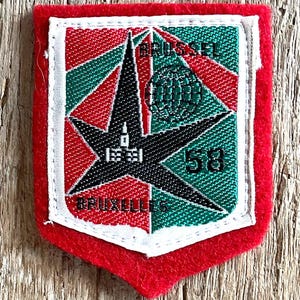 Op de afbeelding: Een vintage patch met een rode rand en een schildvormig ontwerp. De patch heeft een zwarte ster, rode en groene geometrische patronen en de tekst "BRUSSELS 58". De patch is waarschijnlijk een souvenir of een herdenkingsartikel.
