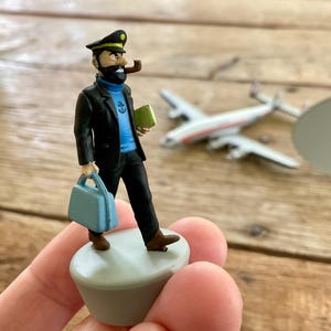 K&ouml;nnte beinhalten: Figur eines Mannes in schwarzer Jacke, blauem Hemd und schwarzer Hose, der eine blaue Aktentasche und ein gr&uuml;nes Buch h&auml;lt. Er tr&auml;gt eine schwarze M&uuml;tze und raucht eine Pfeife. Ein Spielzeugflugzeug ist im Hintergrund. Die Figur steht auf einer kleinen, hellgrauen Plattform.