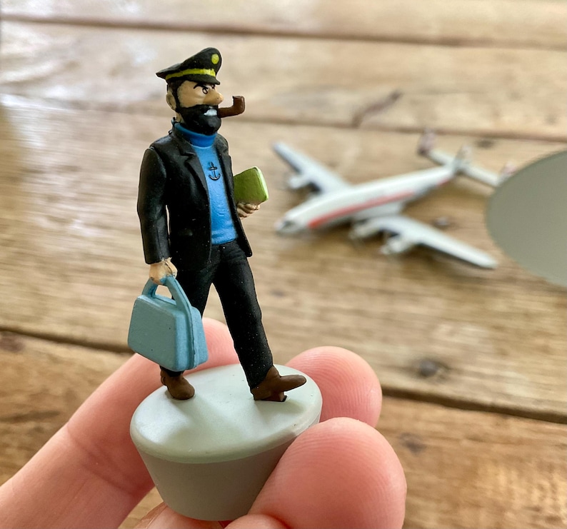 K&ouml;nnte beinhalten: Figur eines Mannes in schwarzer Jacke, blauem Hemd und schwarzer Hose, der eine blaue Aktentasche und ein gr&uuml;nes Buch h&auml;lt. Er tr&auml;gt eine schwarze M&uuml;tze und raucht eine Pfeife. Ein Spielzeugflugzeug ist im Hintergrund. Die Figur steht auf einer kleinen, hellgrauen Plattform.