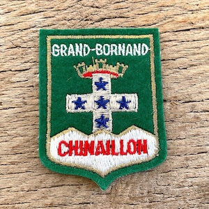 Op de afbeelding: Een groene patch met gouden rand met de tekst "GRAND-BORNAND" en "CHIMAILLON". Het heeft een wit kruis met blauwe sterren en een gouden kroon met rode accenten.
