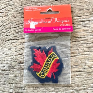 Op de afbeelding: Een Canada souvenir patch met een rood esdoornblad met het woord "CANADA" in geel op een zwarte achtergrond. De patch zit in een doorzichtige plastic zak met een rood-witte kartonnen header met "International Insignia".