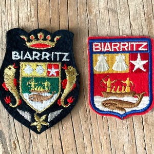 Op de afbeelding: Twee geborduurde patches met de tekst Biarritz. De linker patch is zwart met een gouden kroon, twee gouden zeepaardjes en een centraal schild. De rechter patch is rechthoekig met een rode rand, een boot, een walvis en een witte ster.