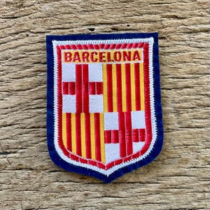 May include: A shield-shaped embroidered patch featuring the Barcelona coat of arms with red and yellow vertical stripes and a red cross on a white background. The word BARCELONA is stitched in red letters at the top, bordered by a dark blue edge.