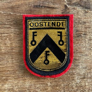Op de afbeelding: Een rechthoekige patch met een rode rand, zwarte bies en een gouden centrum. De patch heeft bovenaan het woord "OOSTENDE", een zwarte chevron en drie zwarte sleutels. De patch is waarschijnlijk een militair of organisatorisch embleem.