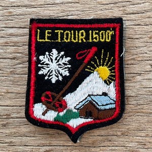 Puede incluir: Parche bordado con fondo negro y borde rojo. El parche presenta el texto "LE TOUR 1500" en amarillo, un copo de nieve blanco, un bastón de esquí, un sol, una montaña y una cabaña con tejado azul.