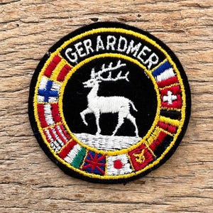 Op de afbeelding: Een ronde geborduurde patch met het woord "GERARDMER" in gele letters. De patch toont een wit hert in het midden, omringd door vlaggen van verschillende landen. De patch heeft een zwarte achtergrond en een gele rand.