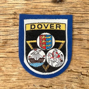 Op de afbeelding: Een blauw en wit embleem met het woord "DOVER" in gouden letters. Het embleem heeft de vorm van een schild met drie cirkelvormige emblemen die een schip, een ridder te paard en een wapenschild afbeelden.