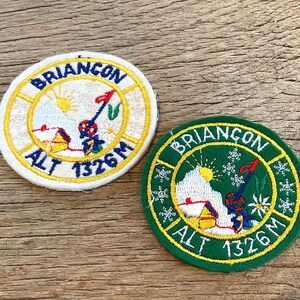 Op de afbeelding: Twee ronde geborduurde patches met de tekst BRIANCON ALT 1326M. De ene patch heeft een witte achtergrond met een gele rand, de andere een groene achtergrond met een gele rand. Beide tonen een berg, een zon en een rode vlag.
