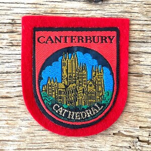 Puede incluir: Un parche rojo con borde negro. Presenta las palabras "CANTERBURY" y "CATHEDRAL". El diseño circular muestra una catedral dorada sobre un cielo azul.