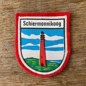 May include: A rectangular patch with a red border and a white stitched edge. The patch features a lighthouse on a green and blue landscape, with the text "Schiermonnikoog" at the top. The background includes blue sky and white clouds.
