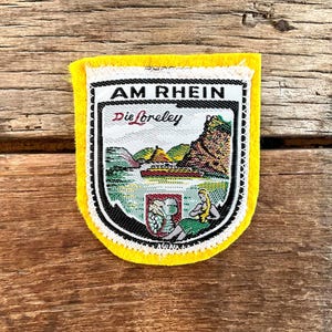 Puede incluir: Un parche amarillo con borde blanco y una imagen detallada de una escena fluvial. El parche presenta el texto "AM RHEIN Die Loreley" y una representación de un paisaje con un barco, montañas y una figura.