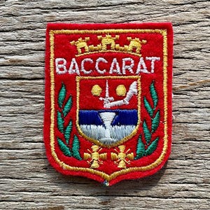 Op de afbeelding: Een rood geborduurd embleem met het woord "BACCARAT" in het wit. Het embleem heeft een wapen met een schildontwerp, gouden accenten en groene en gouden blad details. Het embleem is rechthoekig met afgeronde hoeken.