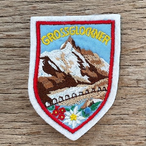 Op de afbeelding: Geborduurde patch met de Grossglockner-berg. De patch heeft een schildvorm met een rode rand en witte achterkant. Het ontwerp toont een berglandschap met het woord "GROSSGLOCKNER" in geel en kleurrijke bloemen.