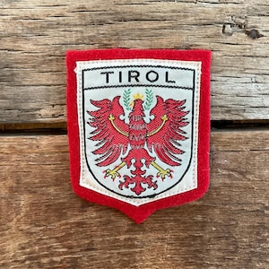 Op de afbeelding: Een rood-wit geborduurde patch met het woord "TIROL" bovenaan. De patch heeft een rode rand en een centraal ontwerp van een rode adelaar met uitgespreide vleugels. De adelaar staat tegen een witte achtergrond.