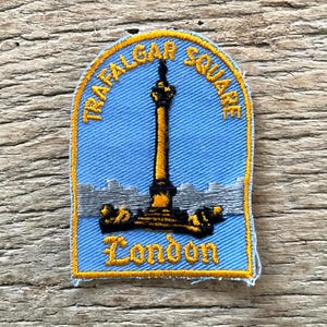 Op de afbeelding: Geborduurde patch met de Nelson's Column op Trafalgar Square, Londen. De patch heeft een lichtblauwe achtergrond met gouden en zwarte details, en de woorden "Trafalgar Square" en "London".