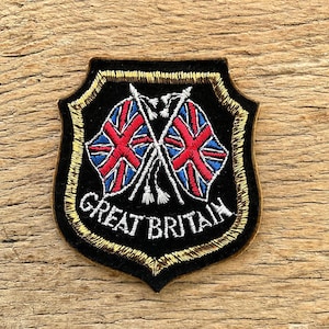 Op de afbeelding: Zwart geborduurd schildvormig embleem met een gouden rand. Het toont twee gekruiste Union Jack-vlaggen en de witte woorden "GREAT BRITAIN". Een patriottisch embleem.