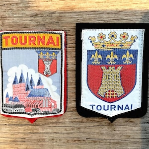 以下が含まれることがあります： 「TOURNAI」の文字が入った2つの刺繍パッチ。1つは赤い縁取りで、青い尖塔のある建物が描かれています。もう1つは黒い縁取りで、王冠とユリの紋章が描かれています。