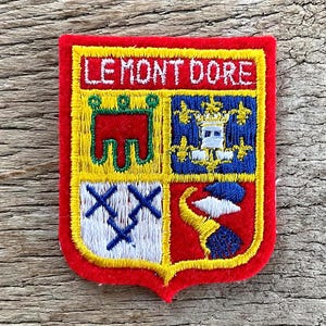 Op de afbeelding: Een rechthoekige patch met een rode rand en een gele binnenkant. De patch heeft een wapenontwerp met de tekst "LE MONT DORE" bovenaan. Het wapen is verdeeld in vier secties met verschillende kleurrijke symbolen.
