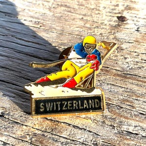 以下が含まれることがあります： 黄色いヘルメット、青いジャケット、黄色のズボンを着たスキーヤーが雪の斜面を滑っているヴィンテージピン。 ピンは金色の縁取りで、「SWITZERLAND」の文字が黒色で書かれています。