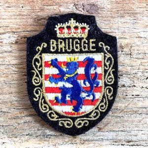 Op de afbeelding: Een zwart geborduurd embleem met het woord "BRUGGE" en een schildontwerp. Het schild toont een blauwe leeuw op een rood-wit gestreepte achtergrond, bekroond met een gouden kroon. Gouden filigraan omlijst het embleem.