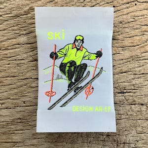 Op de afbeelding: Wit stoffen label met een skiër illustratie. Het woord "SKI" staat in neongeel boven de skiër. De skiër draagt een neongele jas en een zwarte broek. De skistokken zijn oranje. De tekst "DESIGN AR-EF" staat eronder.