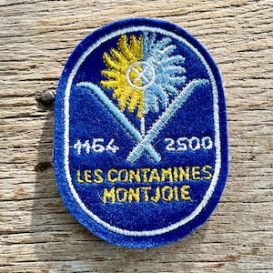 Op de afbeelding: Een blauwe geborduurde patch met een witte rand. De patch heeft een geel en blauw bloemenontwerp, gekruiste ski's en de tekst "LES CONTAMINES MONTJOIE". De nummers "4164" en "2500" zijn ook aanwezig.