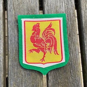 Op de afbeelding: Een schildvormige patch met een felgroene vilten rand rondom een geel veld met een witte omlijning. In het midden van het gele veld staat een rode geborduurde haan in profiel, staand op één poot met uitgespreide staartveren.