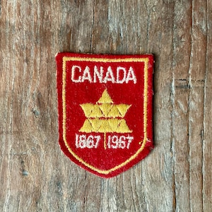 Op de afbeelding: Een rood en goudkleurig Canada-patch met het woord 'CANADA' bovenaan en de jaartallen '1867 1967' onderaan. De patch heeft een gouden sterontwerp in het midden en is omrand met gouden stiksels.
