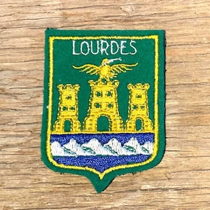 May include: A green embroidered patch with the word "LOURDES" at the top. Below the text is a gold eagle, and three gold castle towers. The bottom of the patch has a blue and white wave design.