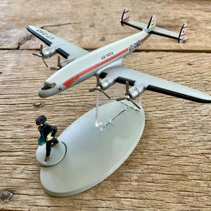 K&ouml;nnte beinhalten: Ein silbernes Modellflugzeug mit der Aufschrift "Air India" und "VT-DAD" auf dem Rumpf. Das Flugzeug hat rote und schwarze Akzente und ist auf einem grauen St&auml;nder montiert. Eine kleine Figur rennt auf dem St&auml;nder.