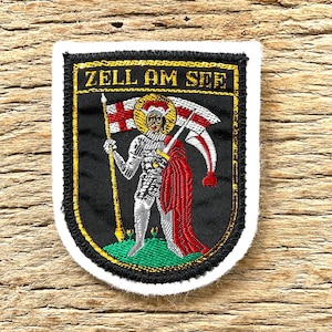 Op de afbeelding: Geborduurde patch met de tekst "ZELL AM SEE" in goud. De patch toont een ridder met een vlag en speer, met een rode mantel. De achtergrond is zwart met een gouden rand en witte rand.