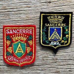 Op de afbeelding: Twee geborduurde patches met het woord "Sancerre". Eén is rood met gouden en groene details, de andere zwart met gouden, blauwe en groene details. Beide hebben een wapenontwerp.