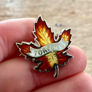 Op de afbeelding: Een esdoornbladbroche met een witte banner met de tekst "TORONTO". De broche heeft een zilveren rand en is gevuld met gele, oranje en rode emaille. Een souvenir of verzamelobject.