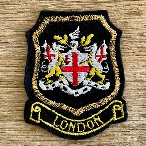 Op de afbeelding: Een zwart geborduurd embleem met een gouden rand en het woord "LONDON" in gouden letters. Het embleem toont een wapen met een rood kruis, witte en gele details en twee leeuwen.